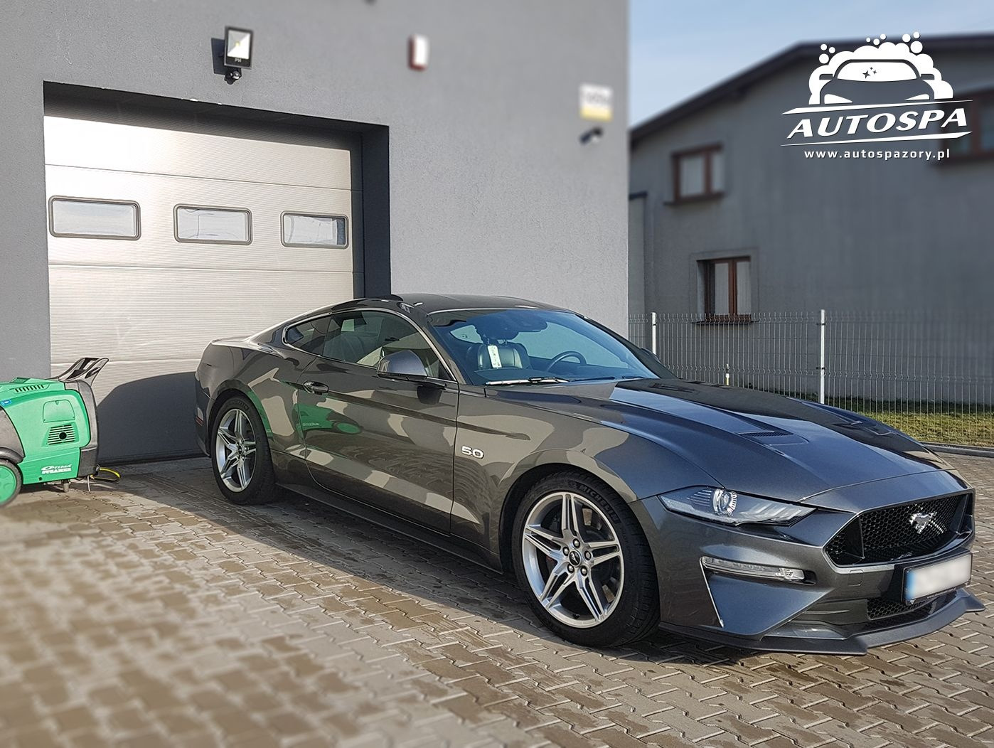 Szary Ford Mustang 5.0 po myciu, zaparkowany przed garażem z widoczną myjką ciśnieniową oraz logo Autospa w prawym górnym rogu.