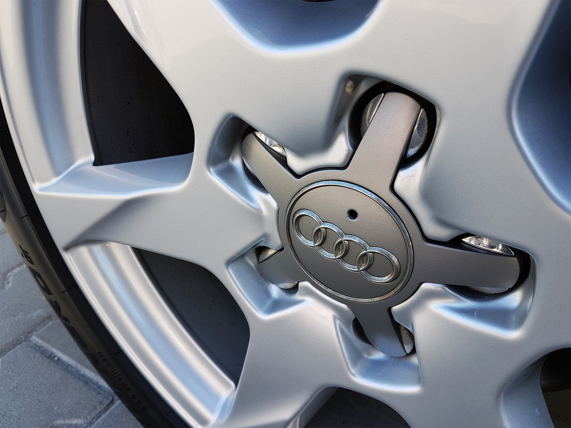 Szczegółowe ujęcie srebrnej felgi aluminiowej z logo Audi, widoczny fragment opony i kostki brukowej w tle.