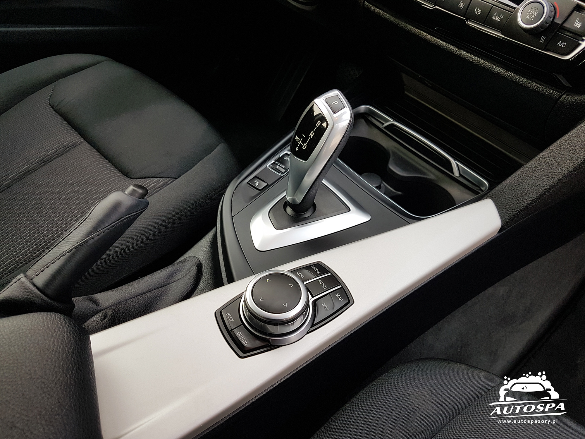 Wnętrze samochodu po detailingu: czysta konsola środkowa z dźwignią zmiany biegów ustawioną w pozycji parkowania, widoczne fotele i logo firmy detailingowej w rogu zdjęcia.