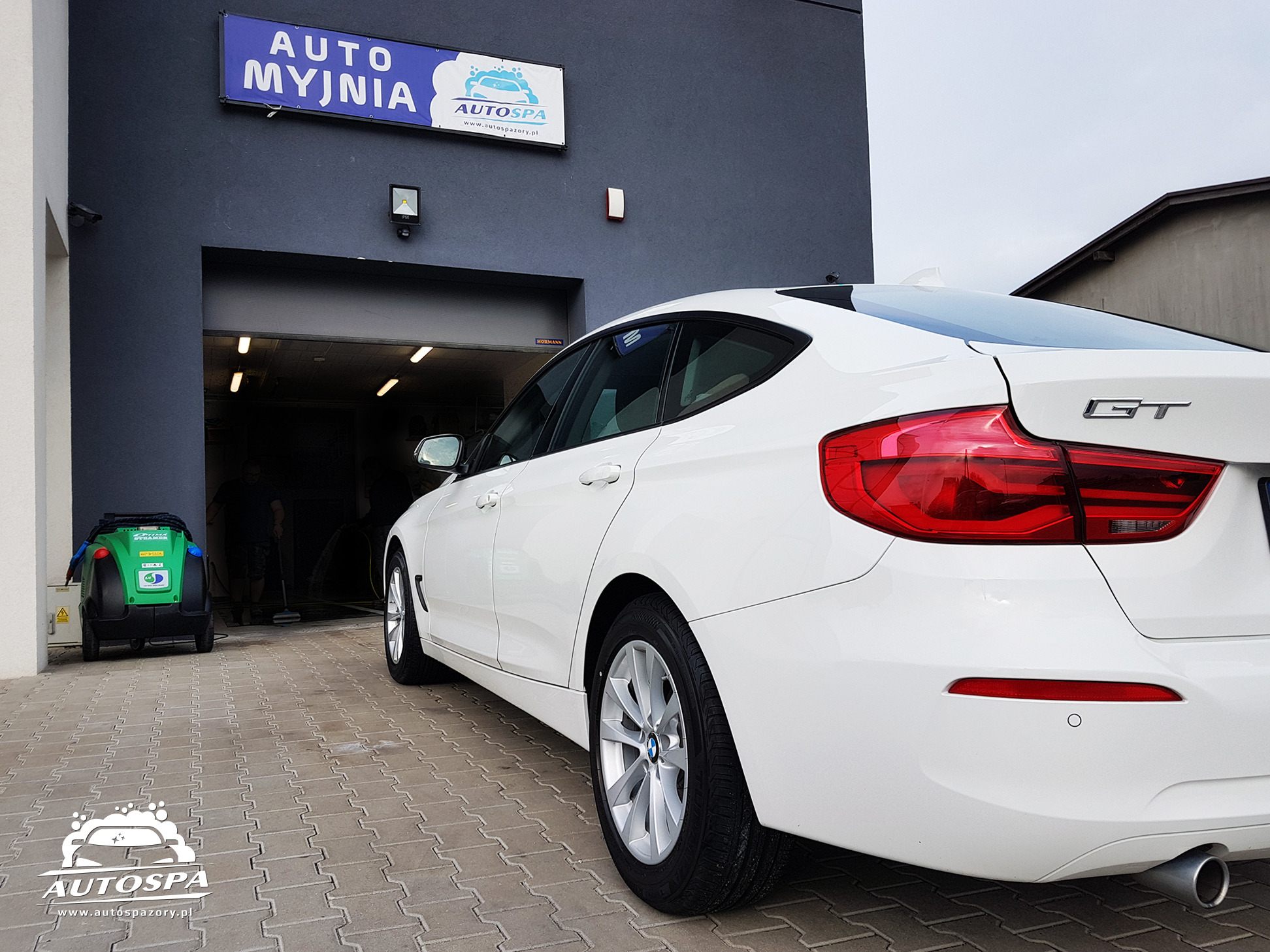 Białe BMW GT zaparkowane przed wjazdem do myjni samochodowej Auto Spa, widoczny sprzęt do czyszczenia w tle.