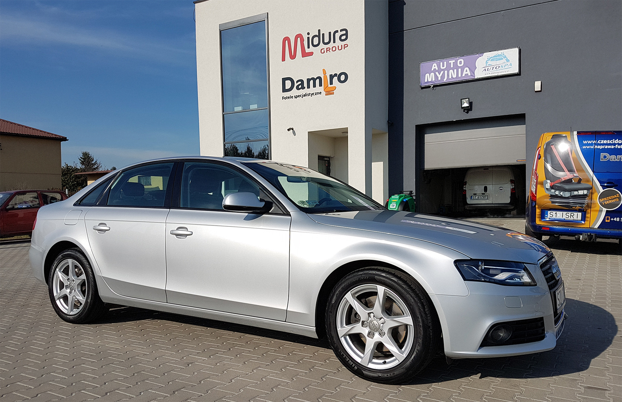Srebrny samochód osobowy zaparkowany na tle budynku z szyldami firmy Midura Group Damro oraz Auto Myjnia Auto Spa, obok zaparkowany bus z reklamą foteli samochodowych.