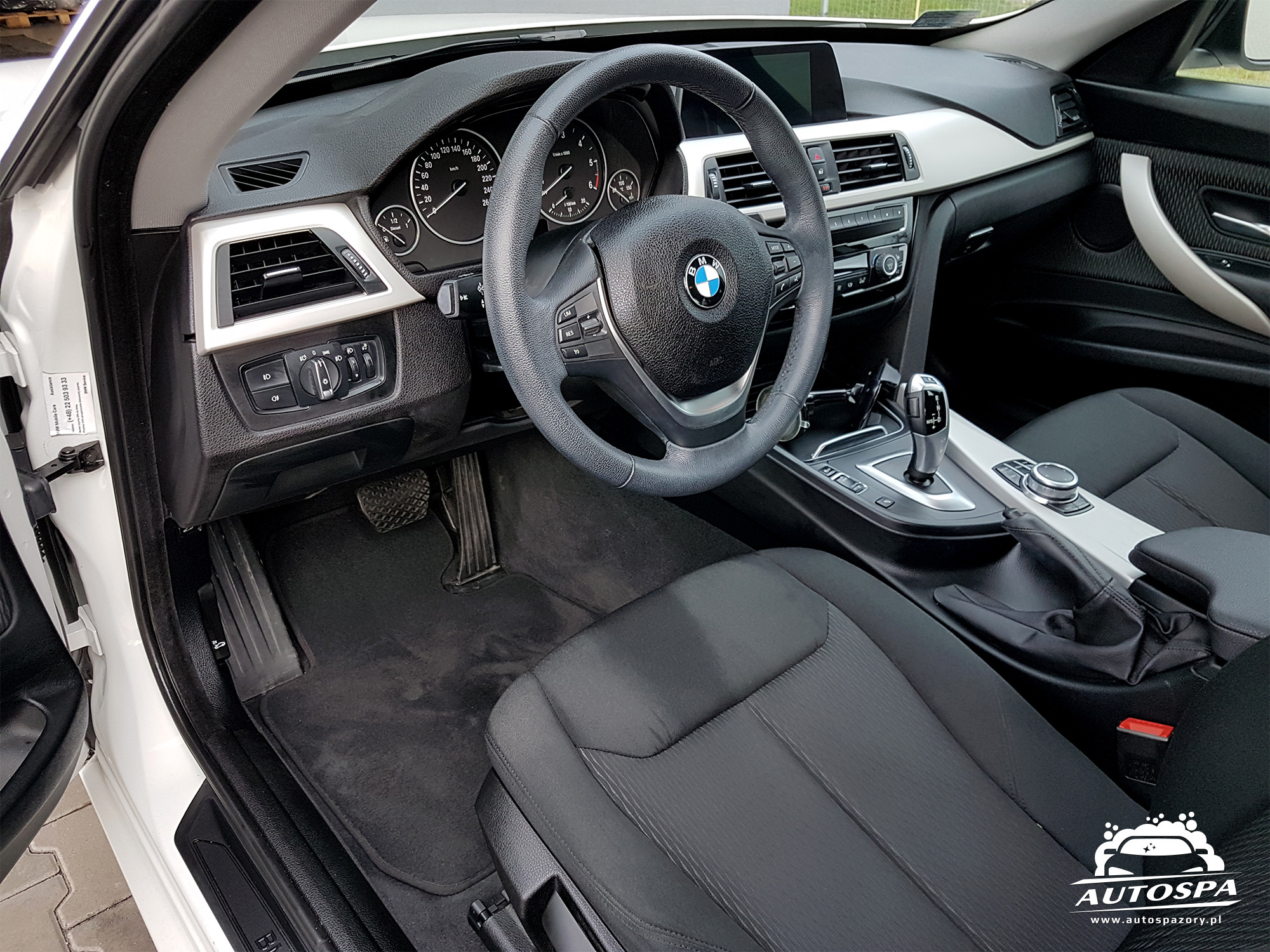 Wnętrze białego samochodu marki BMW po detailingu, widoczna kierownica, deska rozdzielcza, czarne fotele i dywaniki, logo firmy Autospa w prawym dolnym rogu.