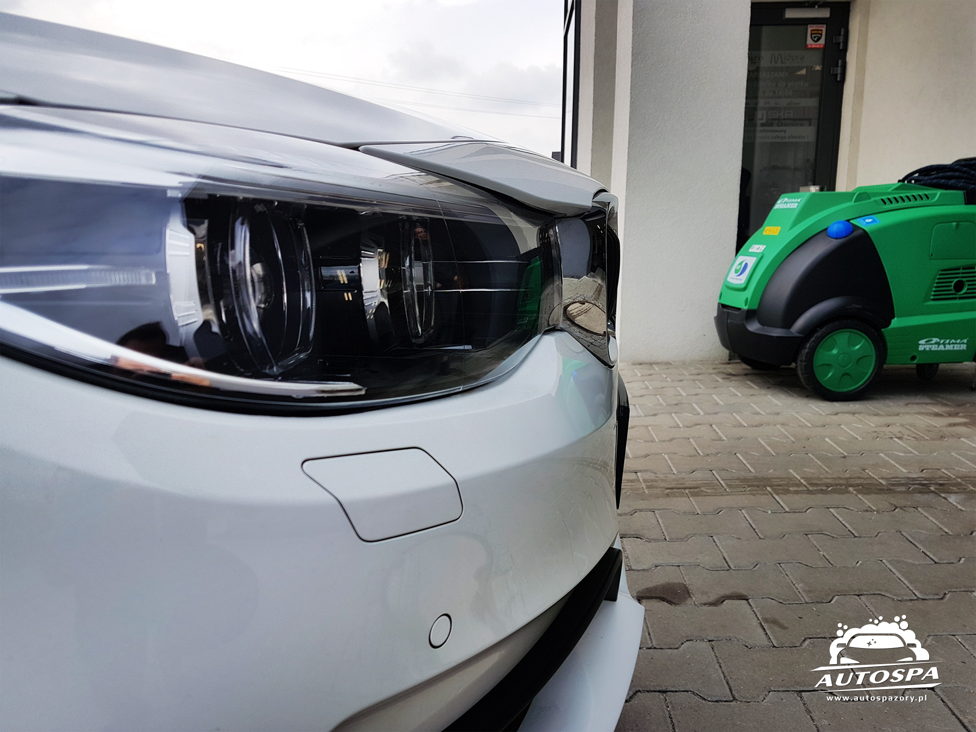 Biały samochód z widocznym reflektorem i czujnikami parkowania, na tle zielonej myjki parowej Optima Steamer i logo AutoSPA na kostce brukowej.