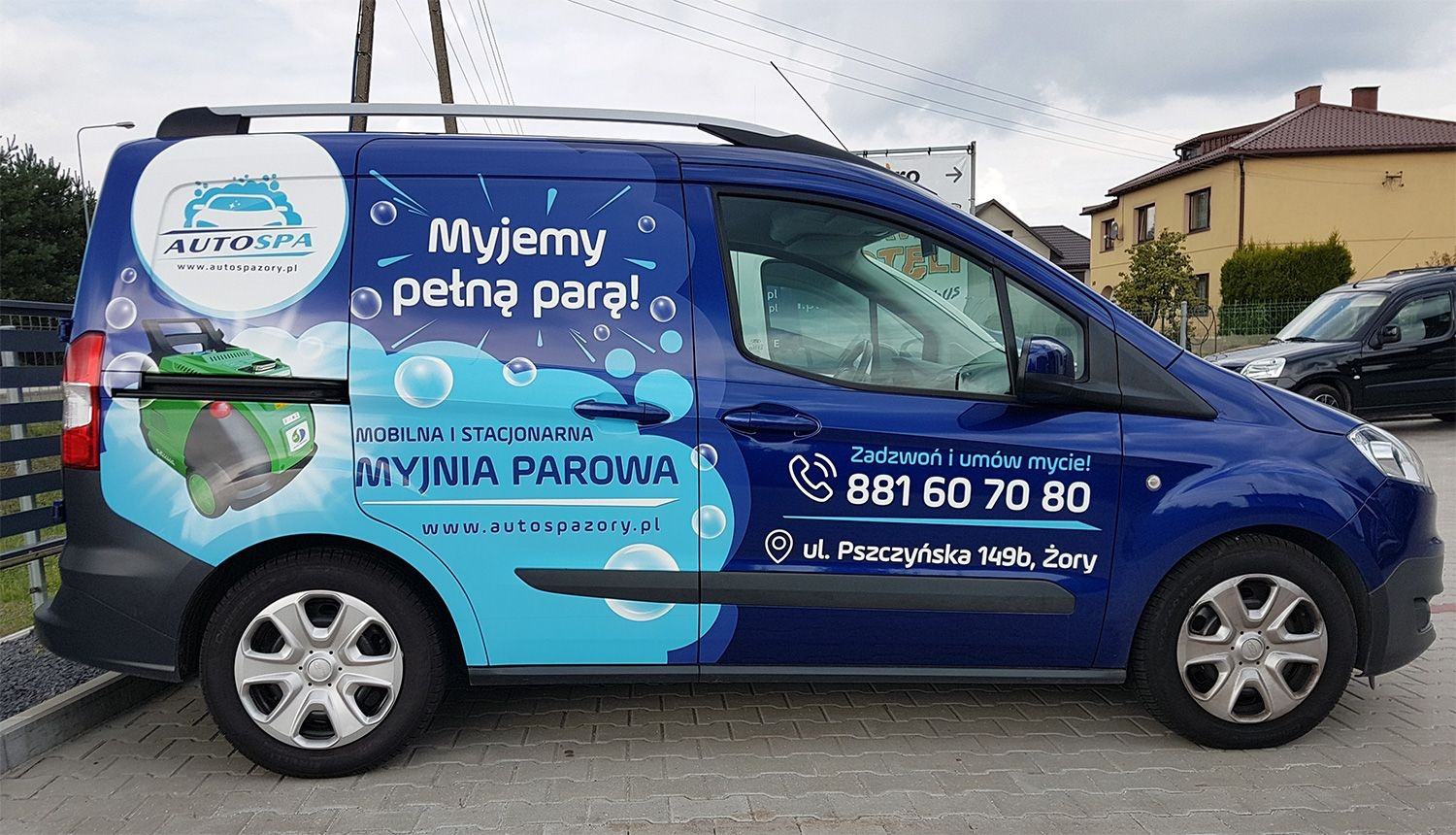 Niebieski van oklejony reklamą mobilnej myjni parowej z numerem telefonu i adresem w Żorach, na tle budynku mieszkalnego.