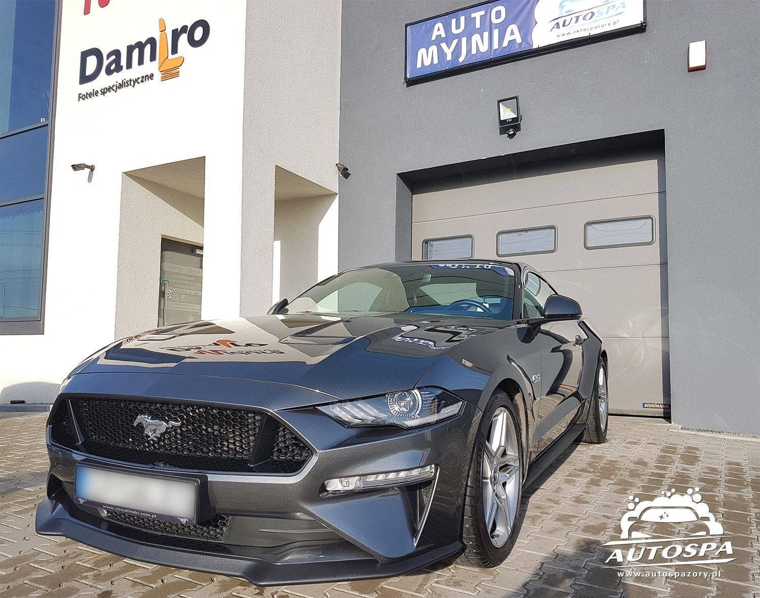 Szary Ford Mustang zaparkowany przed nowoczesną myjnią samochodową z szarymi drzwiami garażowymi i szyldami reklamowymi, kostka brukowa na podłożu.