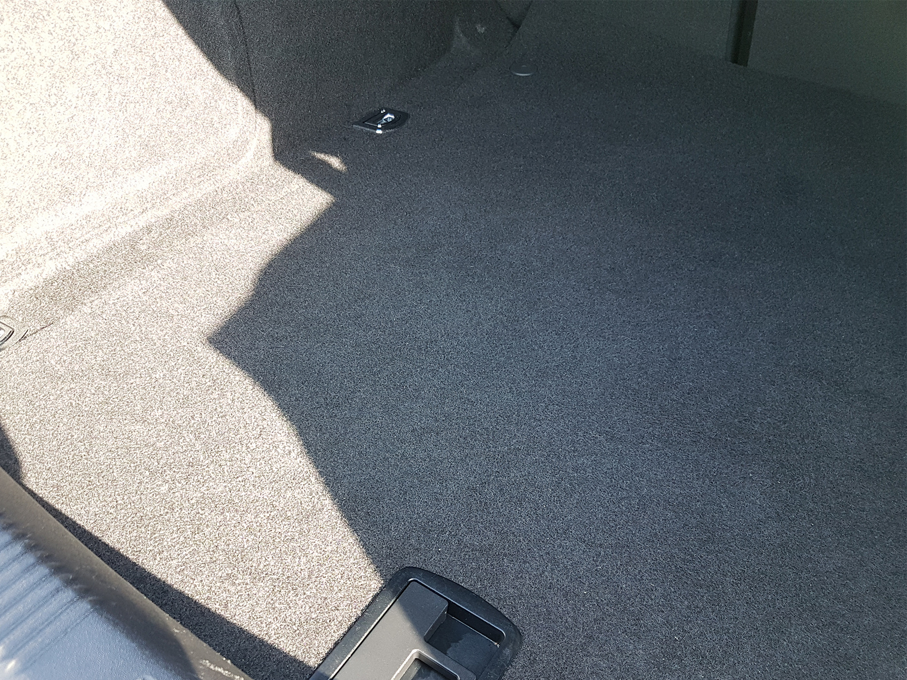 Szara, teksturowana wykładzina bagażnika samochodowego z widocznym mocowaniem i cieniem.