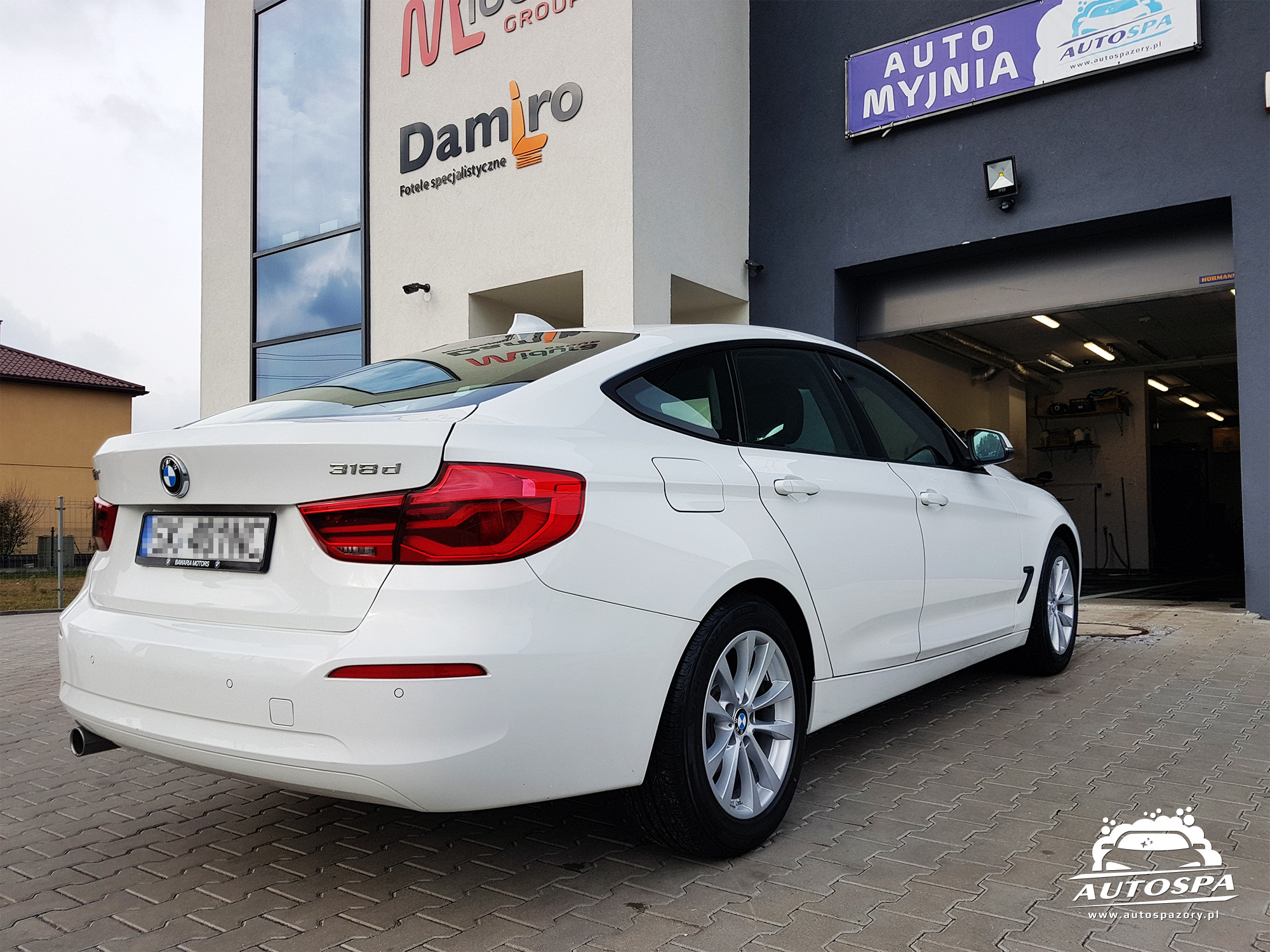 Białe BMW 318d zaparkowane przed budynkiem myjni Auto Spa z widocznym logo firmy na chodniku z kostki brukowej.