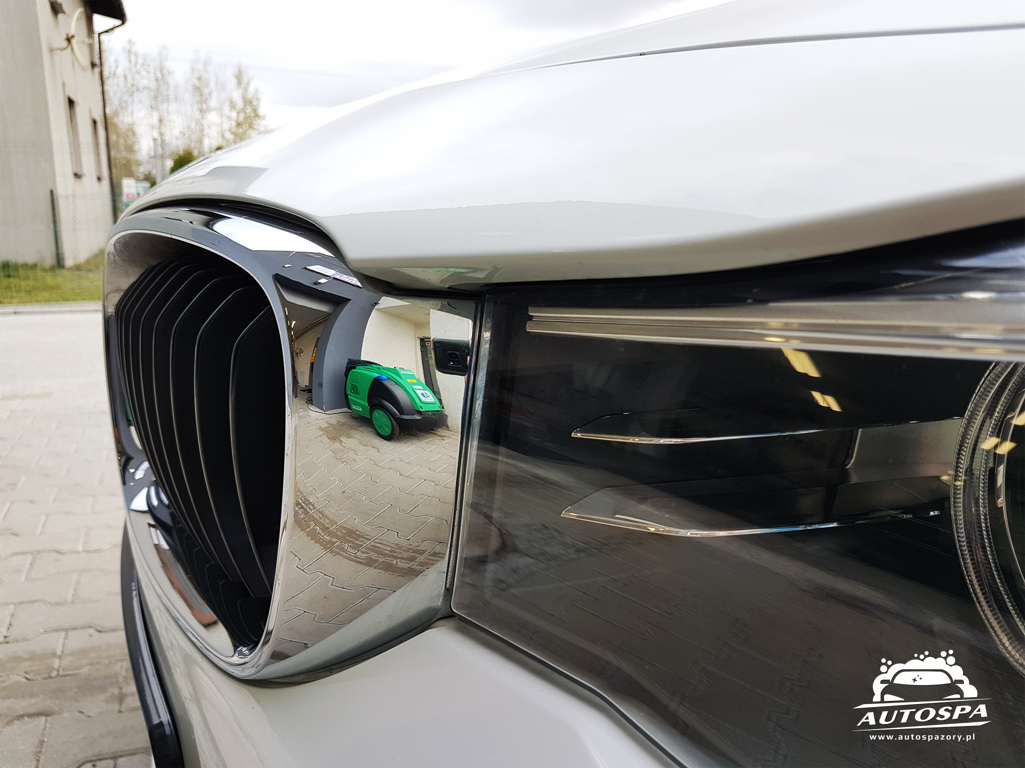 Błyszczący, biały samochód z chromowaną atrapą chłodnicy, w której odbija się zielona myjka ciśnieniowa.