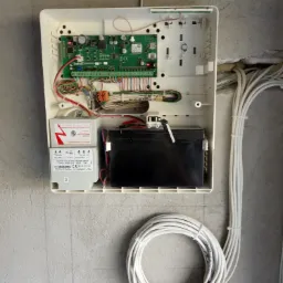 Otwarta obudowa centrali alarmowej z widoczną płytą główną, transformatorem izolującym Noratel i akumulatorem, zamontowana na szarej ścianie. Obok widoczne wiązki przewodów.