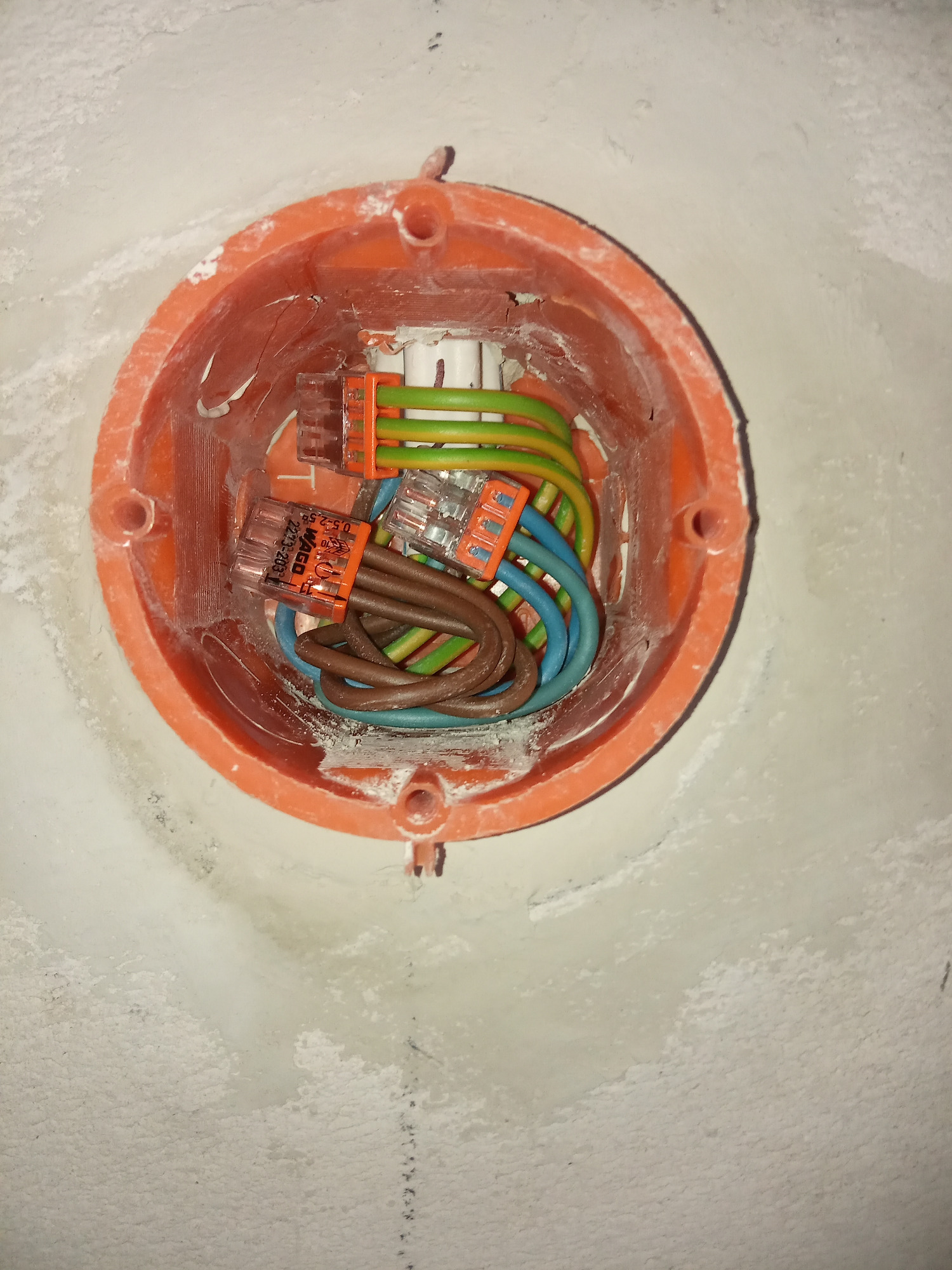 Pomarańczowa puszka instalacyjna w ścianie z widocznymi przewodami elektrycznymi połączonymi złączkami WAGO, widok z góry.