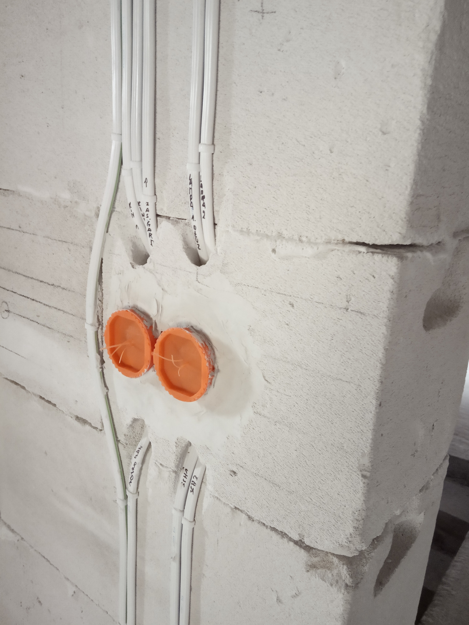 Białe przewody elektryczne prowadzone w ścianie z betonu komórkowego, z widocznymi pomarańczowymi puszkami instalacyjnymi i odręcznymi oznaczeniami na przewodach.