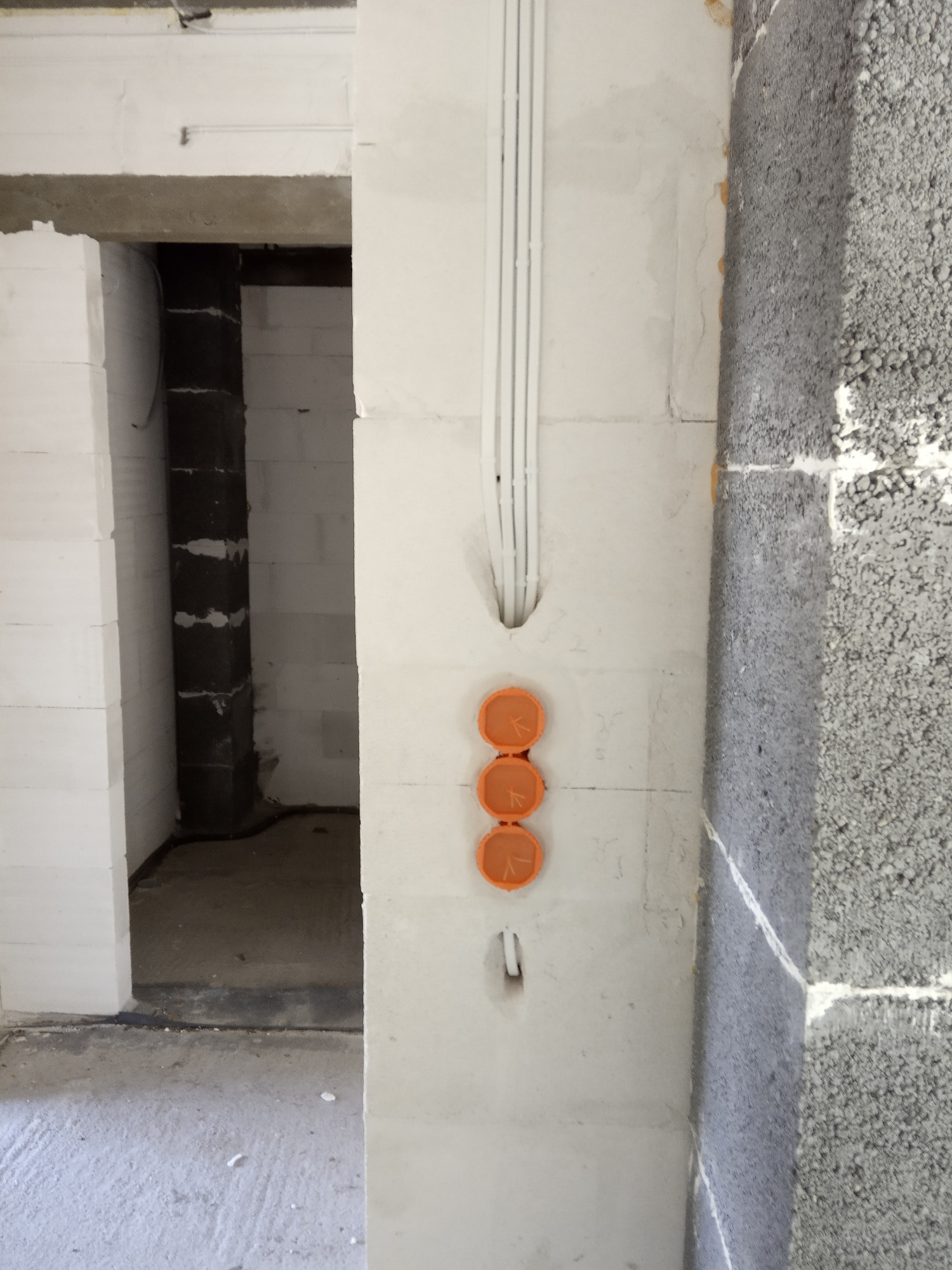 Wewnętrzna ściana budynku w trakcie budowy z trzema pomarańczowymi puszkami instalacyjnymi i białymi rurkami PCV prowadzącymi kable, widok na surowy stan budynku.