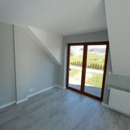 Pomalowane na szaro poddasze z brązowymi drzwiami balkonowymi i widokiem na zielony krajobraz, jasna podłoga imitująca drewno, biały grzejnik.