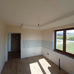 Wnętrze w trakcie wykańczania: ułożona podłoga imitująca drewno, częściowo położona geometryczna glazura na ścianie, widoczne instalacje elektryczne i hydrauliczne, surowe wykończenie okna, drzwi...