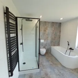 Nowoczesna łazienka z kabiną prysznicową typu walk-in z czarnymi profilami, grzejnikiem drabinkowym w kolorze czarnym, białą wanną i toaletą w zabudowie, wykończona szarą płytką imitującą beton...