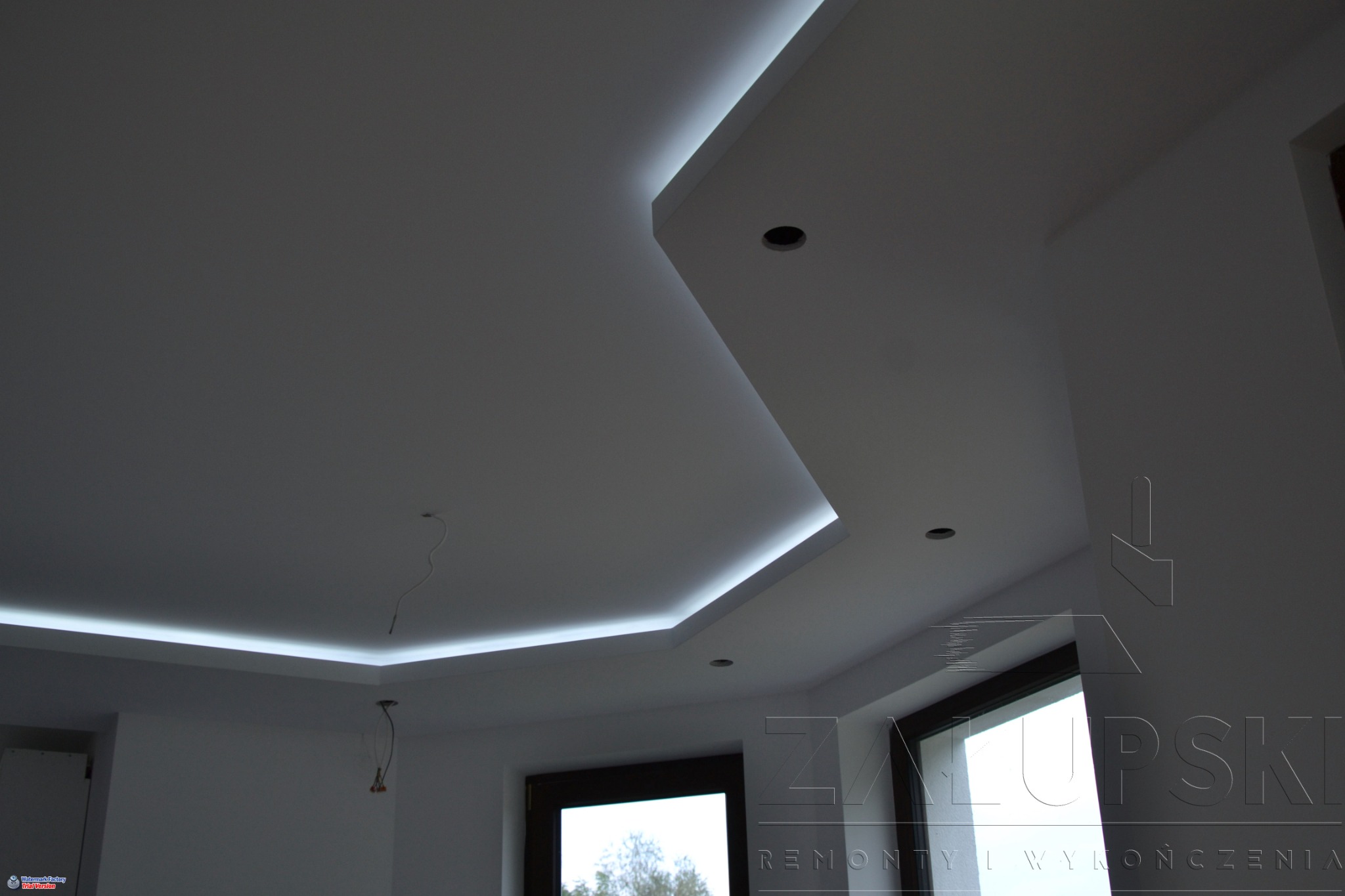 Nowoczesny sufit z podświetleniem LED wzdłuż krawędzi, widoczne otwory na oświetlenie punktowe, fragment okna w tle.