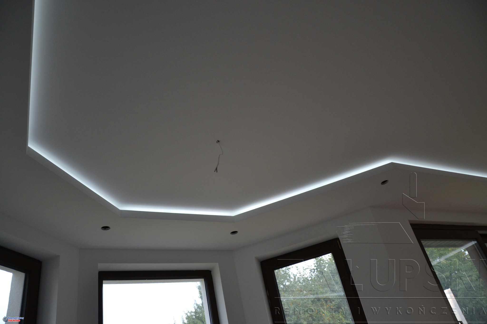 Nowoczesny sufit podwieszany z oświetleniem LED w kształcie litery L, widoczne okna z ciemnymi ramami i wystający kabel.