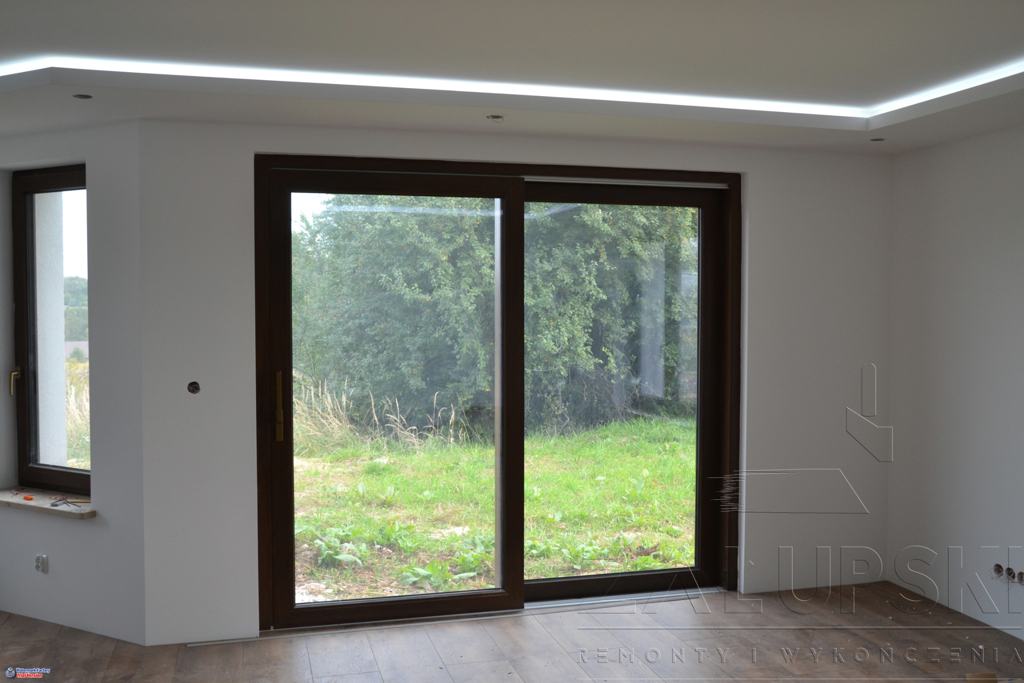 Wykończone wnętrze z dużym przesuwnym oknem tarasowym z brązowymi ramami, widok na zewnątrz na zieleń, białe ściany, oświetlenie LED na suficie, podłoga panelowa, gniazdka elektryczne w ścianie.