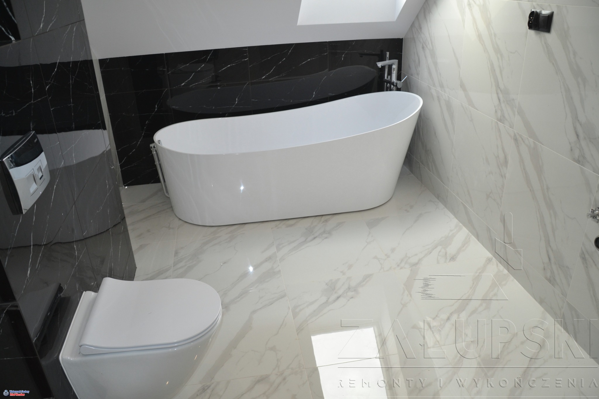 Nowoczesna łazienka na poddaszu z czarno-białymi marmurkowymi płytkami, wolnostojąca biała wanna, podwieszana toaleta i oświetlenie sufitowe.