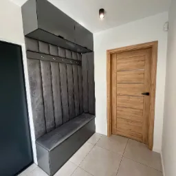 Nowoczesny przedpokój z szarą, tapicerowaną wnęką z wieszakami i siedziskiem, po lewej ciemne drzwi, po prawej drzwi drewniane.