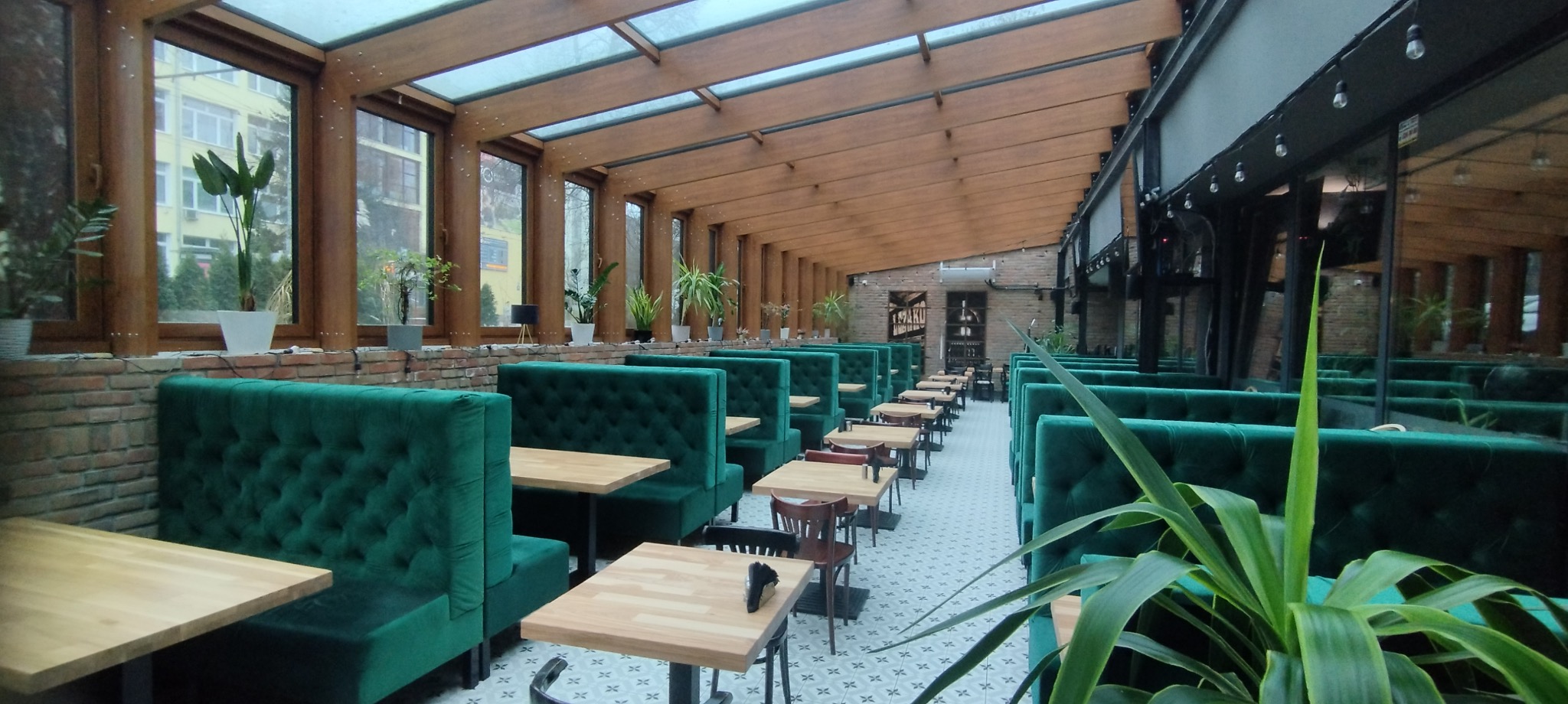 Wnętrze restauracji z rzędami zielonych, pikowanych sof z drewnianymi blatami, przeszklonym dachem i licznymi roślinami doniczkowymi w oknach.