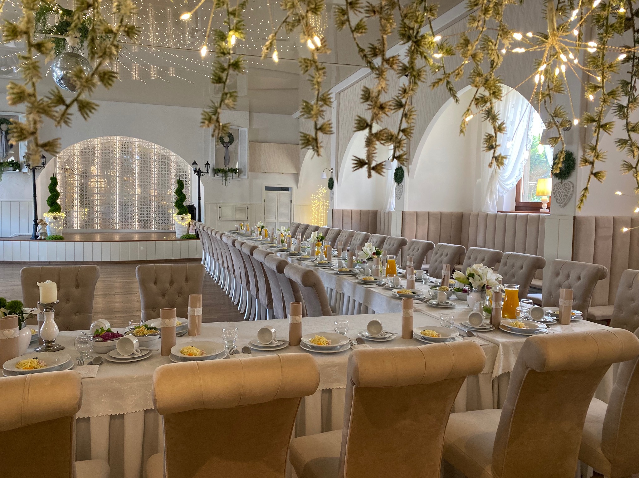 Długi stół weselny nakryty białym obrusem, z beżowymi krzesłami z tapicerowanymi oparciami, udekorowany kwiatami, świecami i zastawą. Na suficie dekoracje świetlne i lustrzana kula.