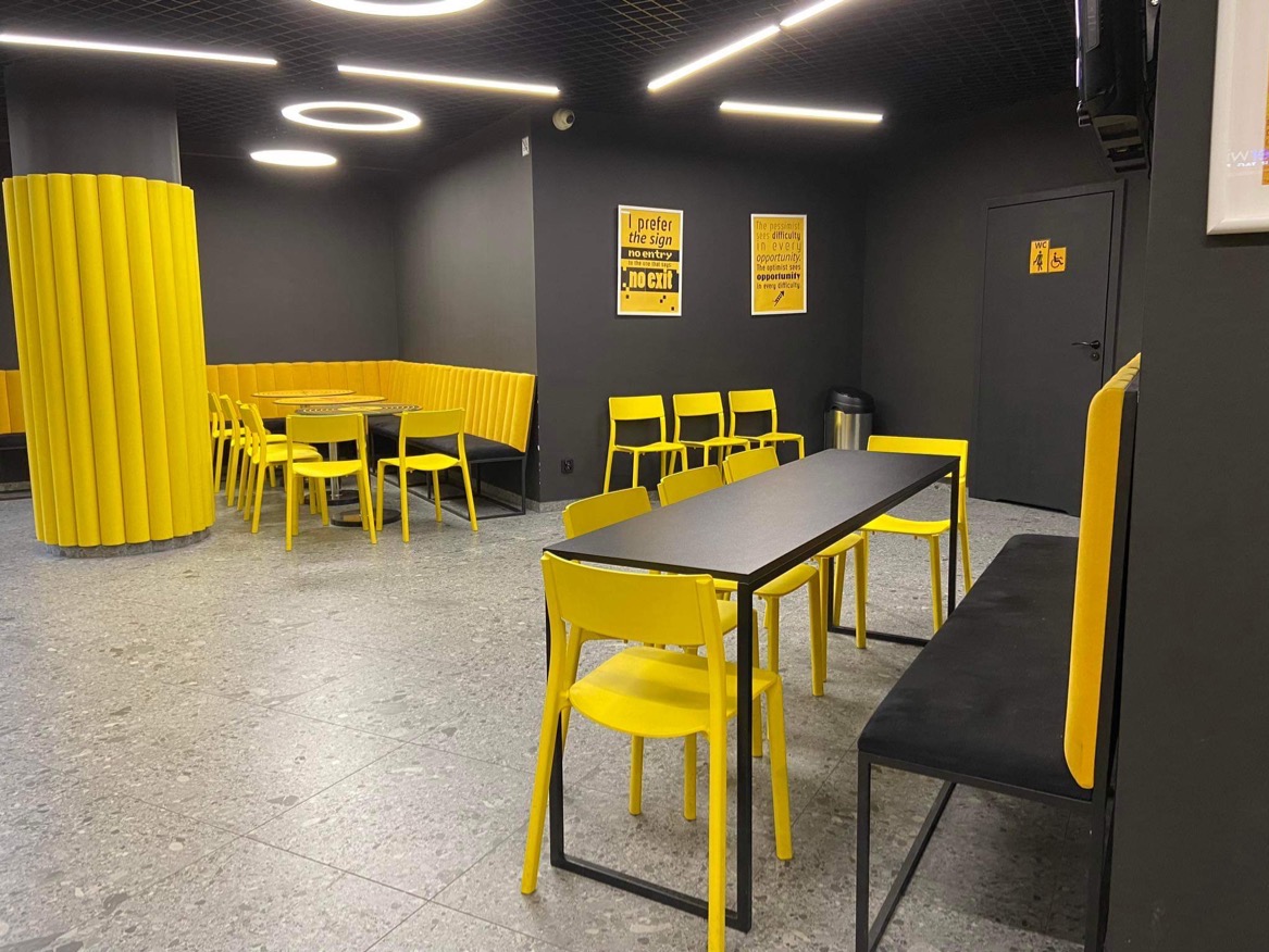 Wnętrze kawiarni lub restauracji z ciemnymi ścianami i podłogą lastryko, ożywione żółtymi krzesłami, obiciami i cylindrycznym elementem dekoracyjnym. Meble tapicerowane z charakterystycznym...