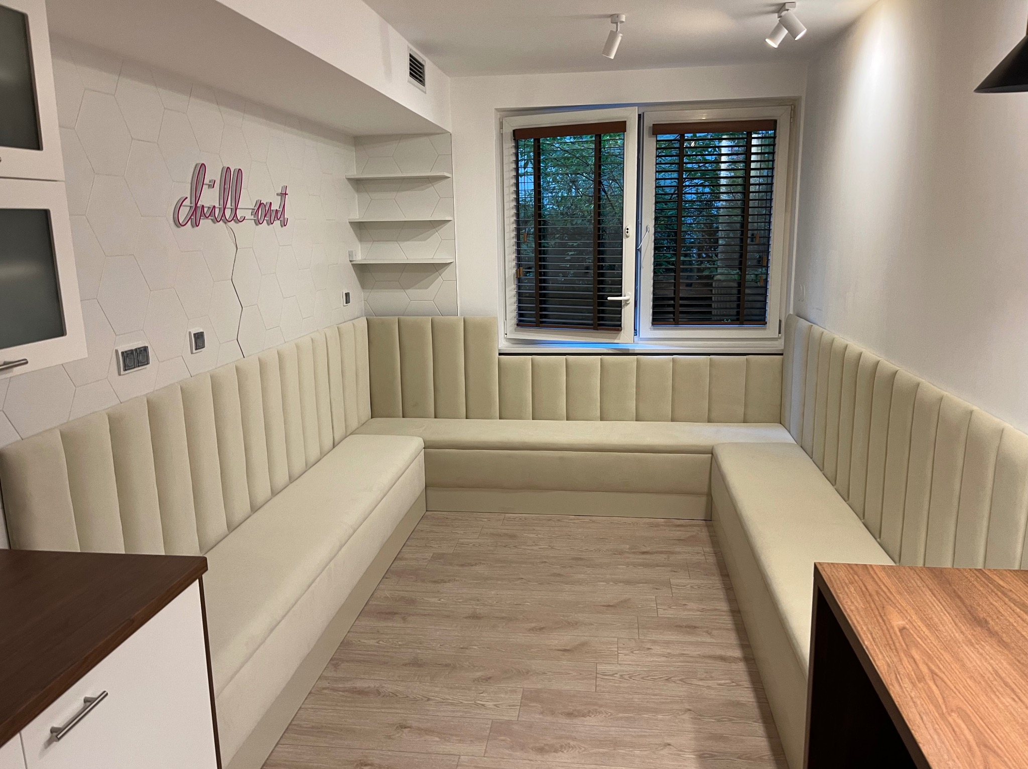 Beżowa, tapicerowana zabudowa siedziska w kształcie litery U pod oknem, z dekoracyjnym napisem 'chill out' na ścianie i drewnianym blatem stołu w rogu.