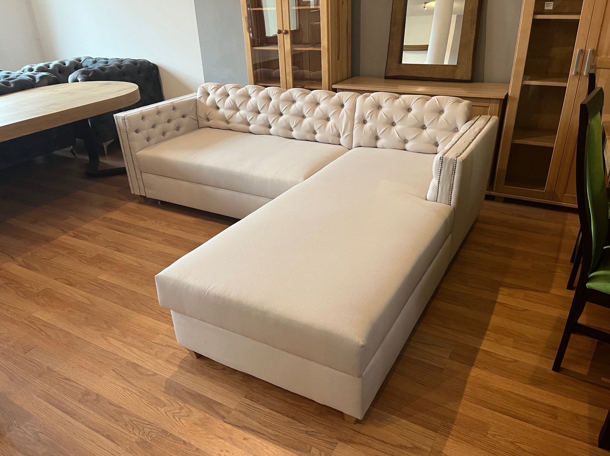 Narożnik tapicerowany w jasnym odcieniu z pikowanym oparciem i podłokietnikami zdobionymi srebrnymi nitami, ustawiony na drewnianej podłodze w aranżacji wnętrza z drewnianymi meblami.