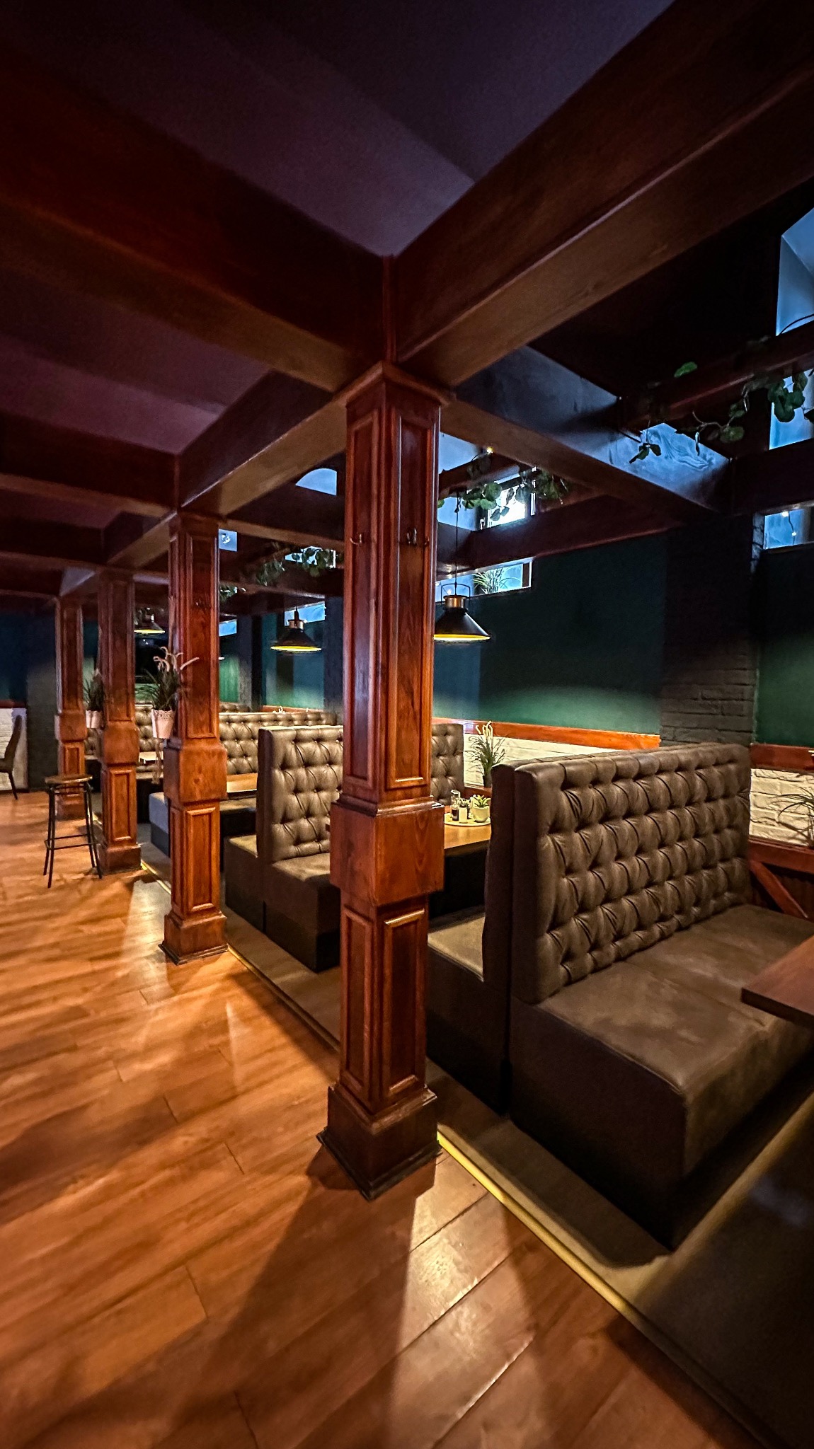 Wnętrze restauracji z rzędami pikowanych, skórzanych kanap w ciemnych odcieniach, oddzielonych drewnianymi kolumnami i belkami stropowymi, oświetlone wiszącymi lampami.