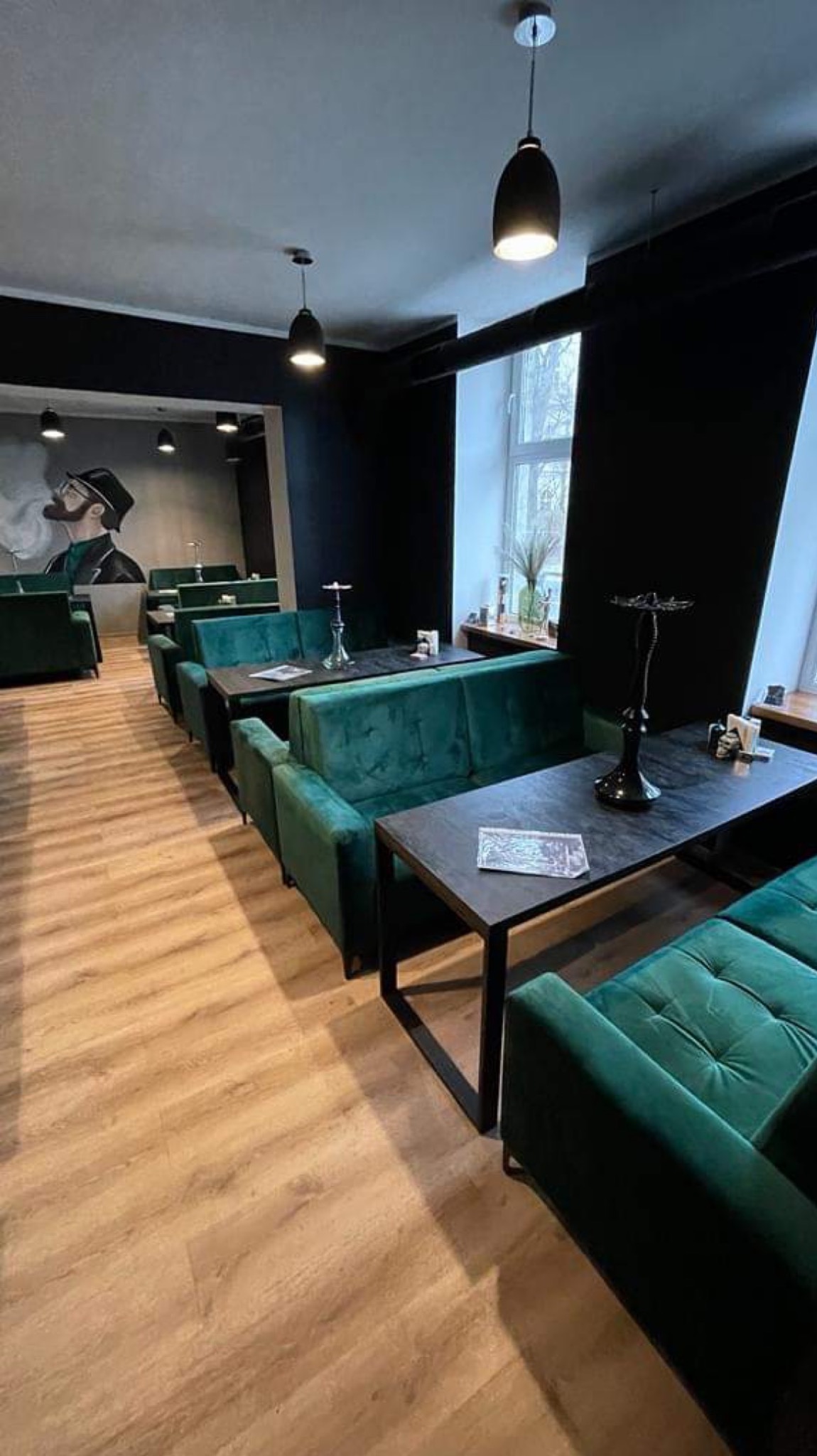 Wnętrze lokalu z rzędem zielonych, tapicerowanych sof i czarnych stolików, oświetlonych wiszącymi lampami; na ścianie mural z palącym mężczyzną.