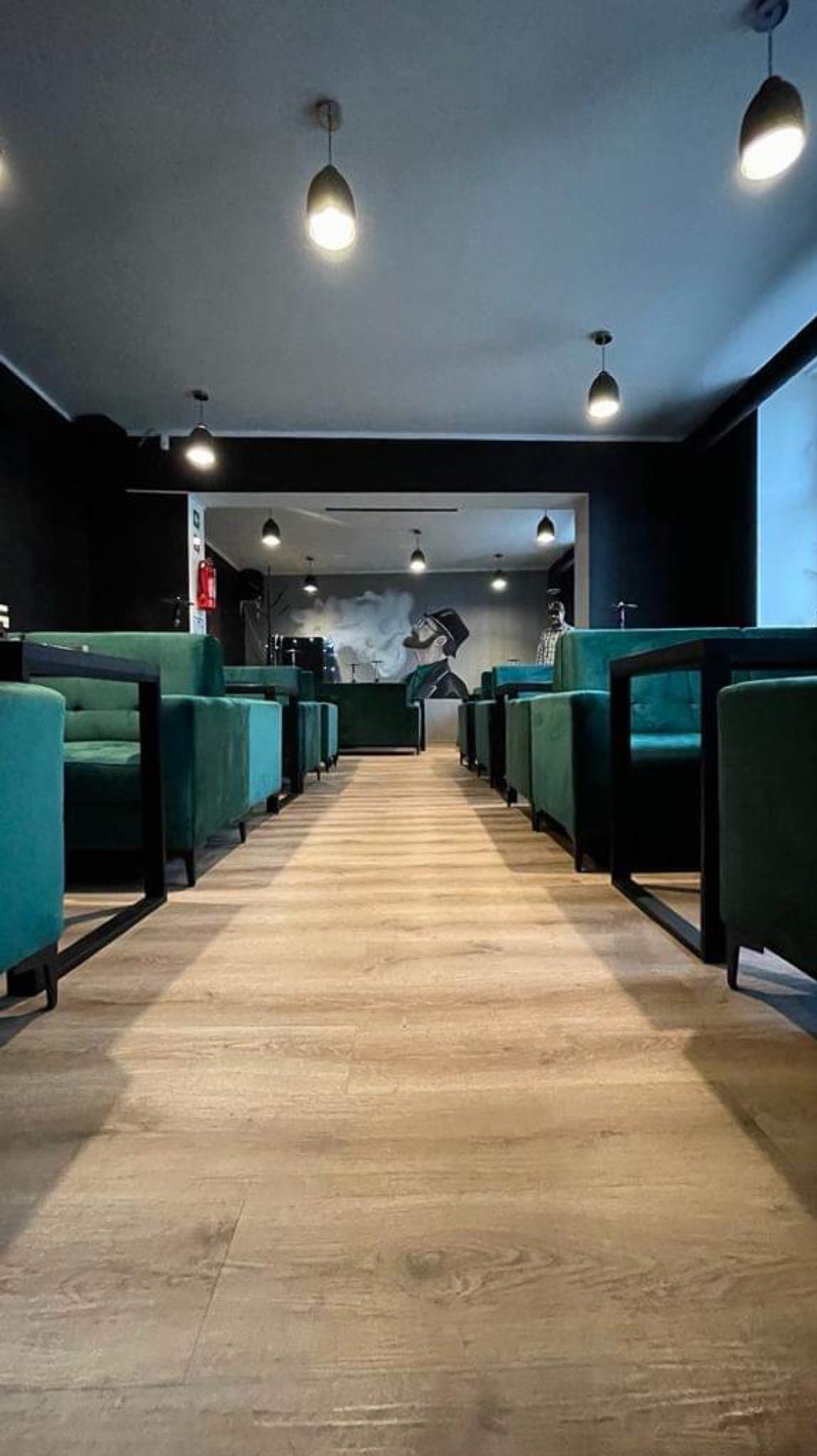 Wnętrze lokalu z rzędem turkusowych, tapicerowanych krzeseł i stolików na tle czarnych ścian i jasnej podłogi, w głębi widoczny mural z postacią palącą fajkę.