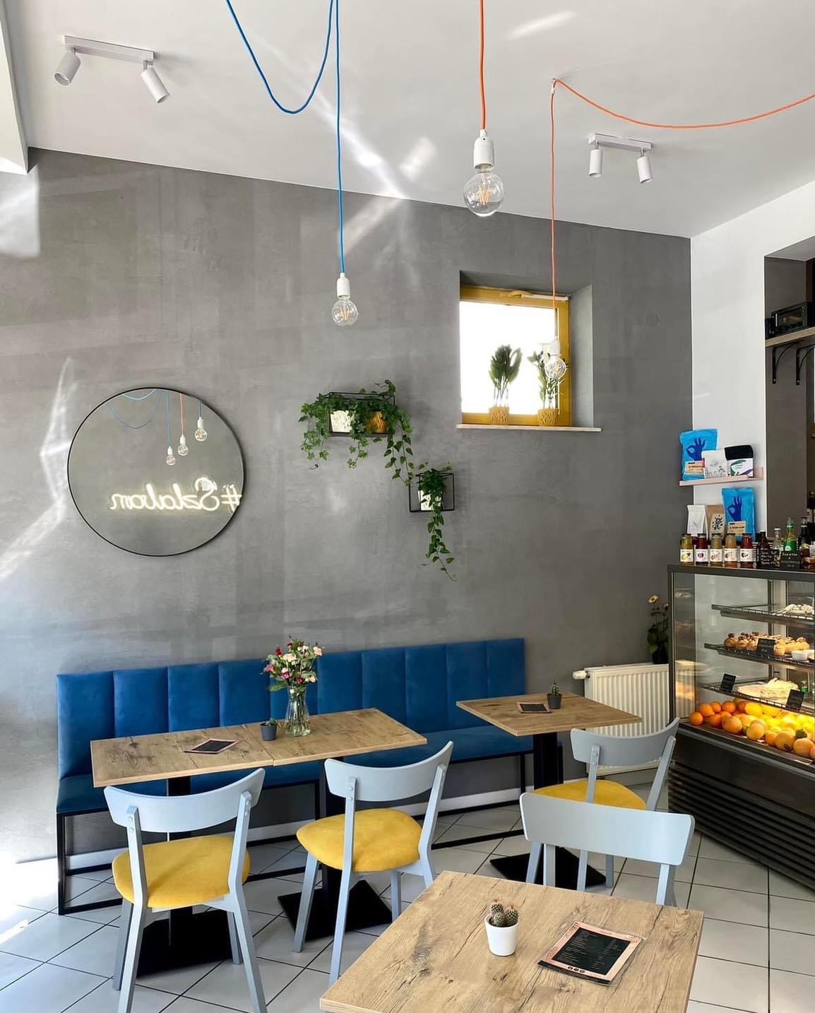 Wnętrze kawiarni z niebieską tapicerowaną ławką, drewnianymi stolikami, szarymi krzesłami z żółtymi siedziskami, betonową ścianą, neonem i roślinami doniczkowymi.