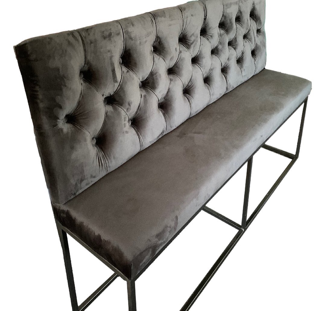 Długa, tapicerowana ławka w kolorze szarym, z pikowanym oparciem i metalową ramą, na białym tle.
