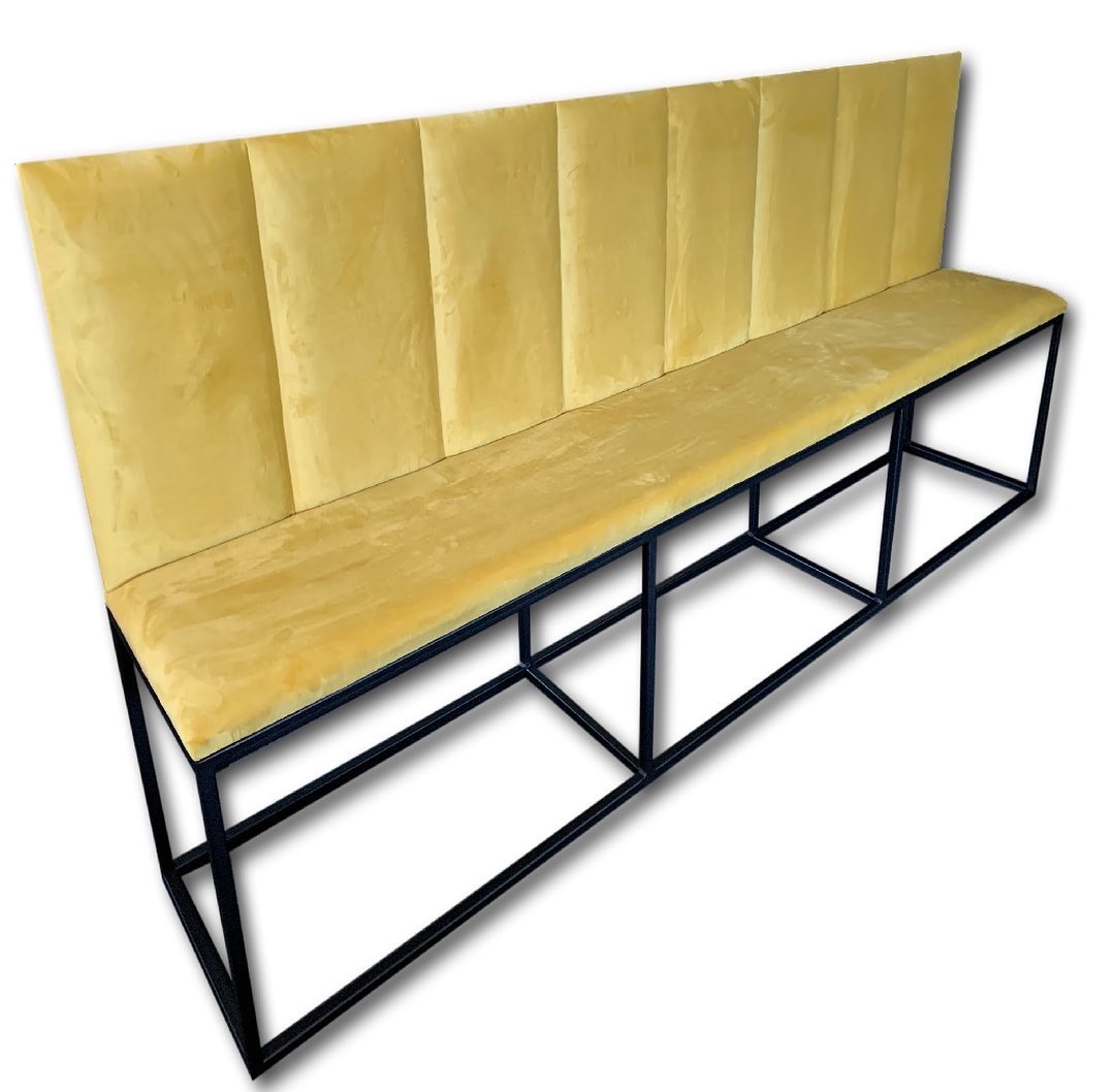 Długa ławka tapicerowana żółtym welurem z wysokim oparciem, na czarnej, metalowej konstrukcji. Nowoczesny design.