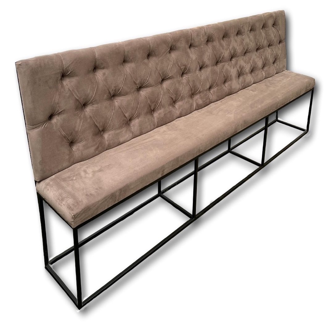 Długa, beżowa ławka tapicerowana z pikowanym oparciem i czarną, metalową podstawą, ustawiona na białym tle.