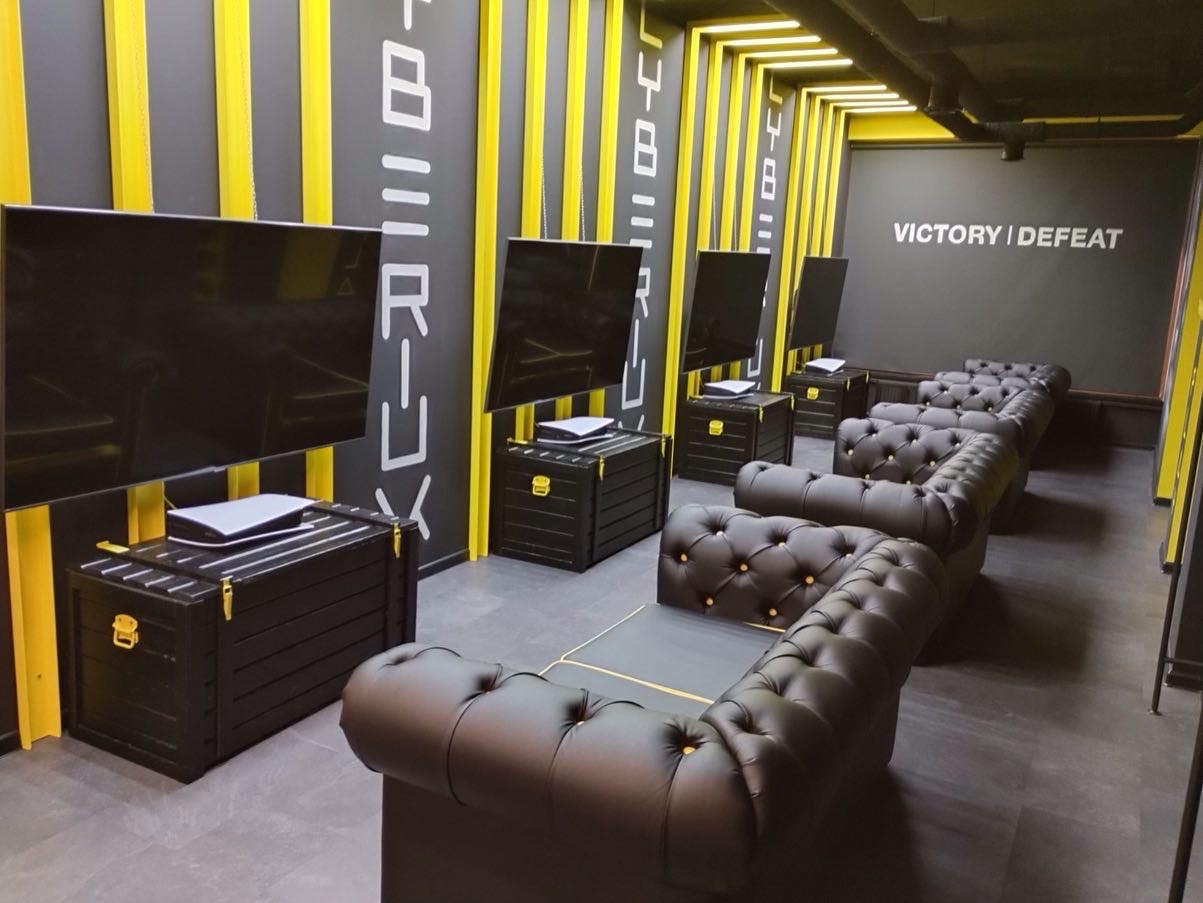 Wnętrze sali gamingowej w czarno-żółtej kolorystyce, z rzędem pikowanych kanap, telewizorami i konsolami na skrzyniach, napis 'Victory / Defeat' na ścianie.