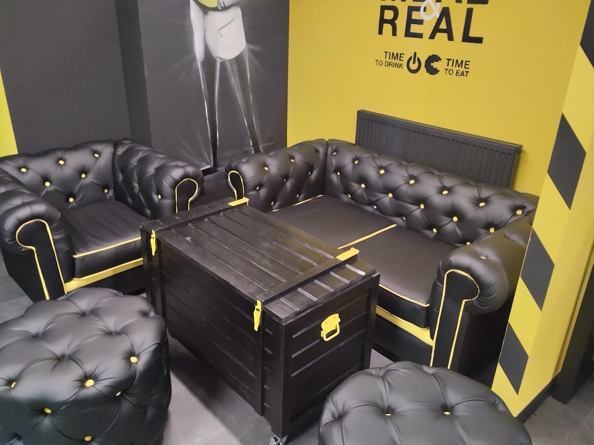 Czarny skórzany narożnik Chesterfield z żółtymi akcentami, pufy i stolik skrzyniowy na kółkach w industrialnym wnętrzu z żółtą ścianą z napisem 'REAL'.
