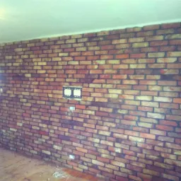 Wnętrze z czerwoną ścianą z cegły klinkierowej, widoczne gniazdka elektryczne i uchwyt na telewizor, podłoga drewniana, częściowo widoczne drzwi.