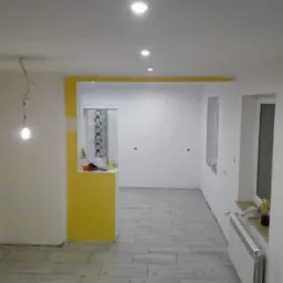 Wnętrze po remoncie: świeżo położona podłoga z płytek imitujących drewno, ż&oacute;łte akcenty malarskie na ścianie, widoczne punkty oświetleniowe w suficie, niedokończone podłączenie lampy wiszącej...