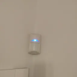 Biały czujnik ruchu alarmu z niebieską diodą LED zamontowany w rogu pomieszczenia na beżowej ścianie.