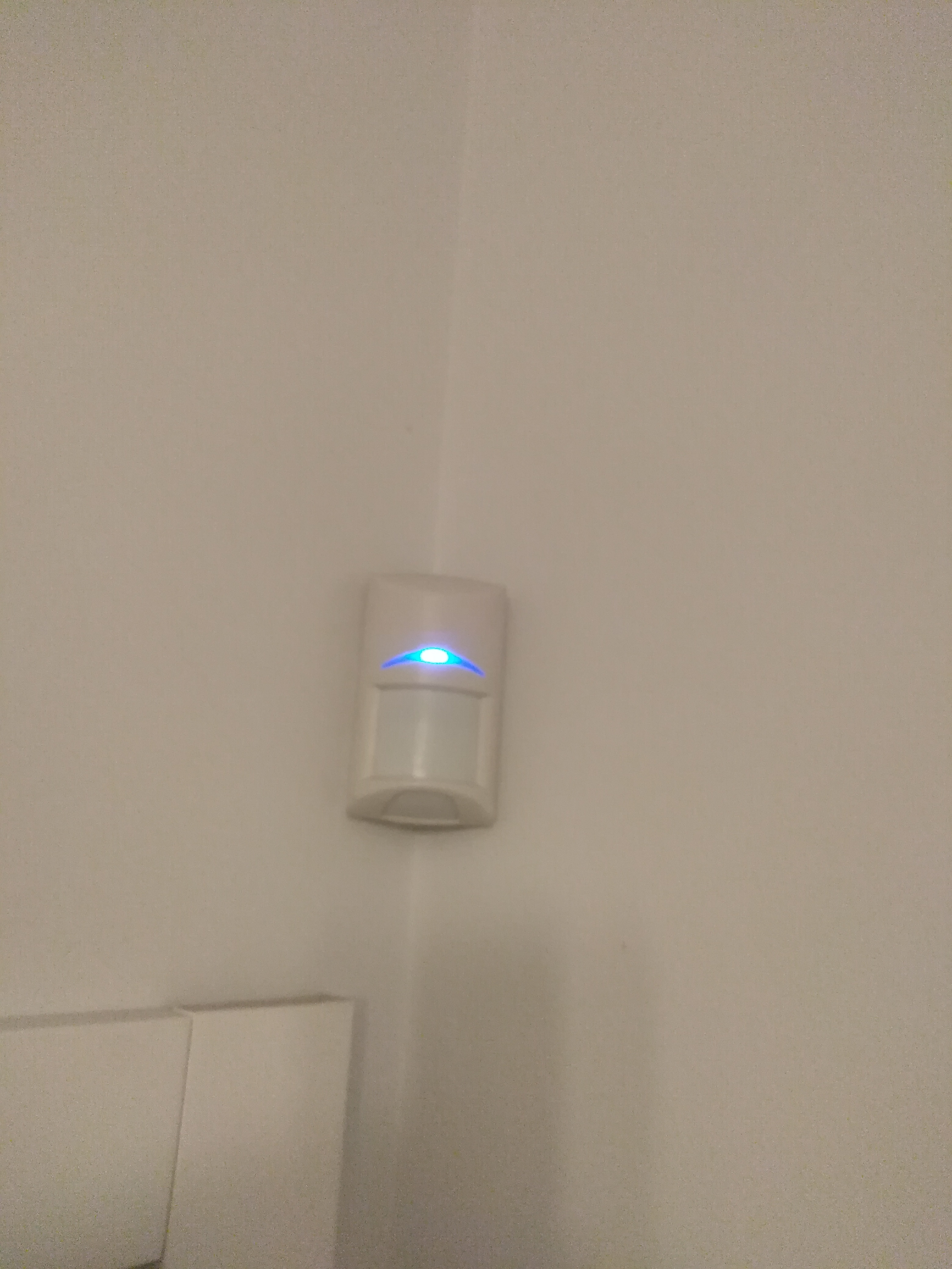 Biały czujnik ruchu alarmu z niebieską diodą LED zamontowany w rogu pomieszczenia na beżowej ścianie.