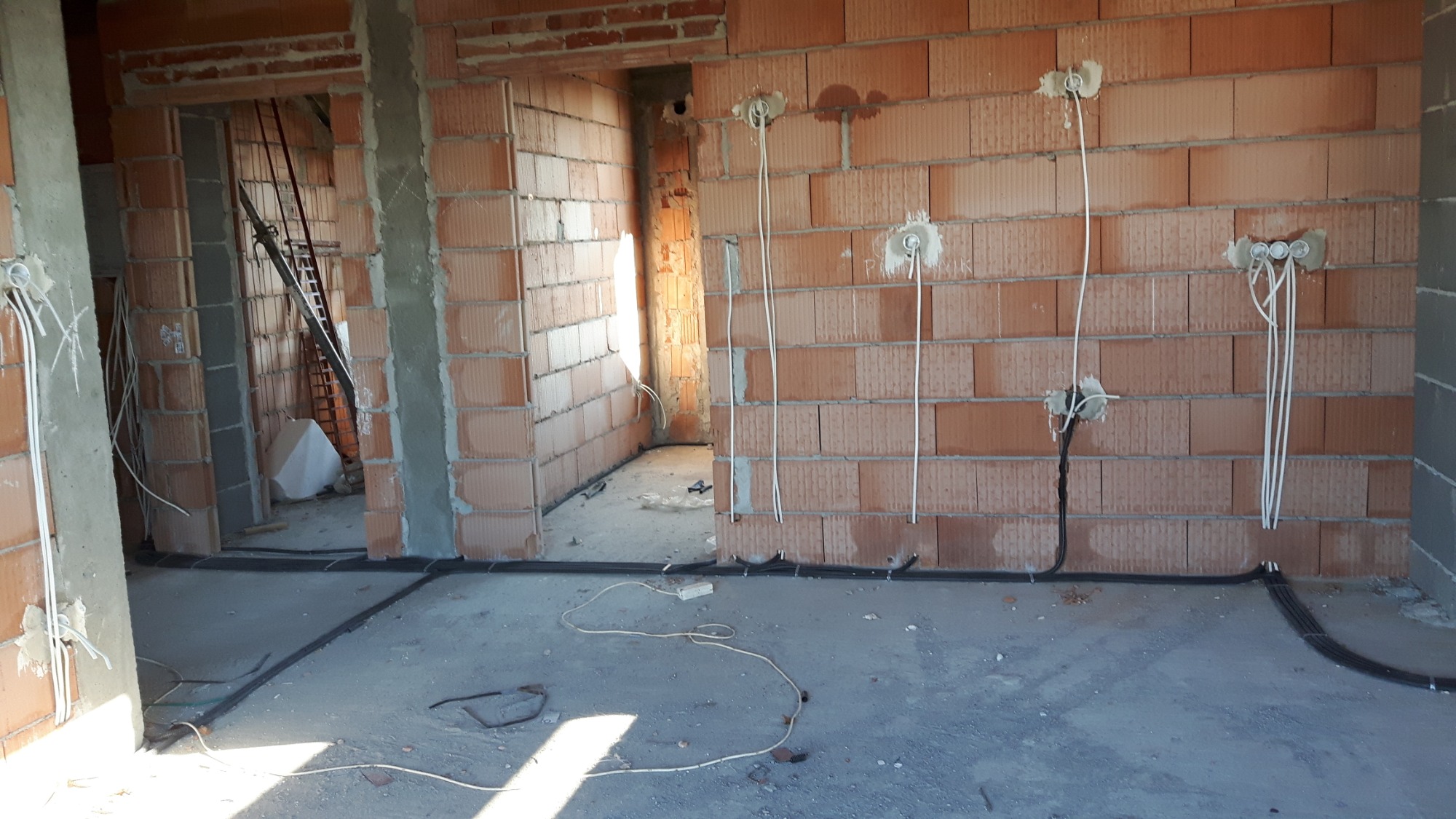 Wnętrze w trakcie budowy z widocznymi przewodami elektrycznymi wystającymi ze ścian z cegły, czarne kable ułożone wzdłuż podłogi.