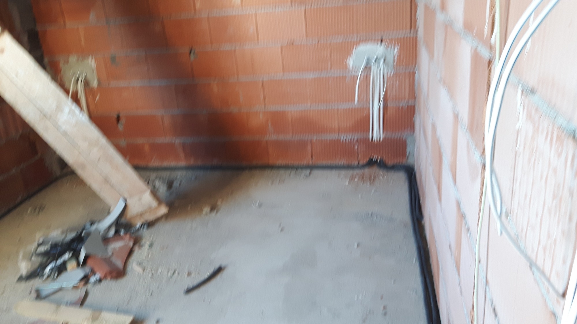 Surowy mur z cegły z wystającymi przewodami elektrycznymi, widoczne kable w peszlu na ścianie i fragmenty gruzu na podłodze.