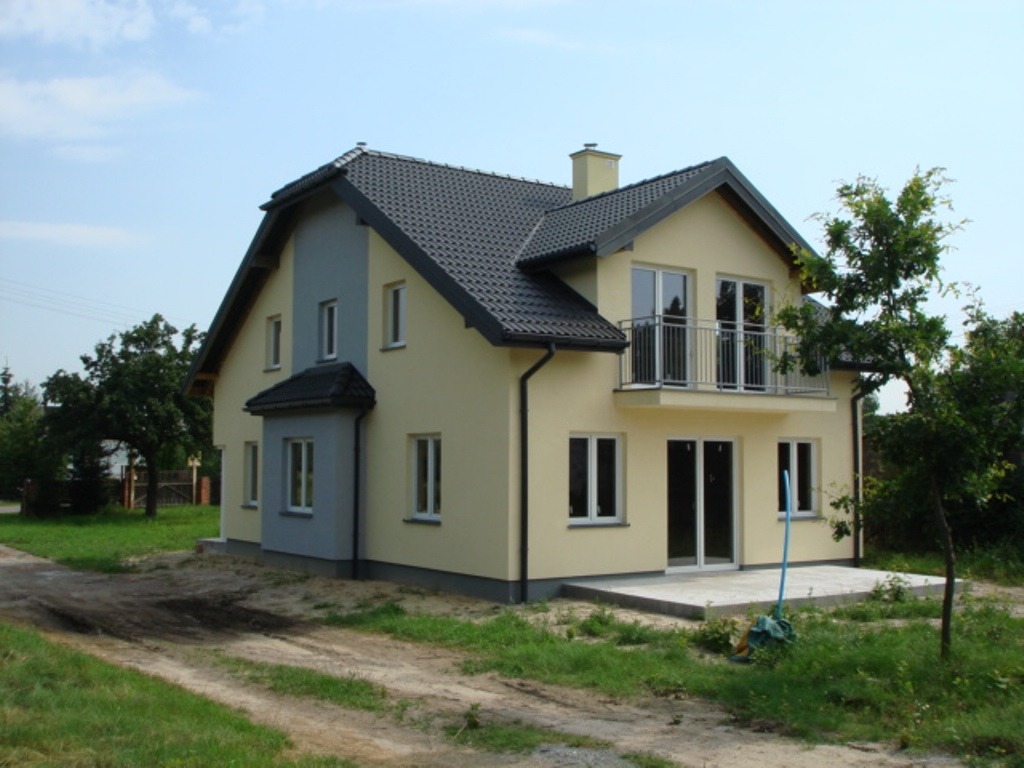 Dwukondygnacyjny dom z beżową elewacją, ciemnoszarą dachówką i balkonem z metalową balustradą, widoczny z perspektywy ścieżki gruntowej.