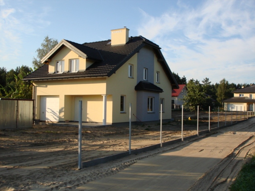 Dwukondygnacyjny dom z dwoma garażami i szarą dachówką, świeżo wykończony, z przygotowanym terenem pod ogrodzenie.