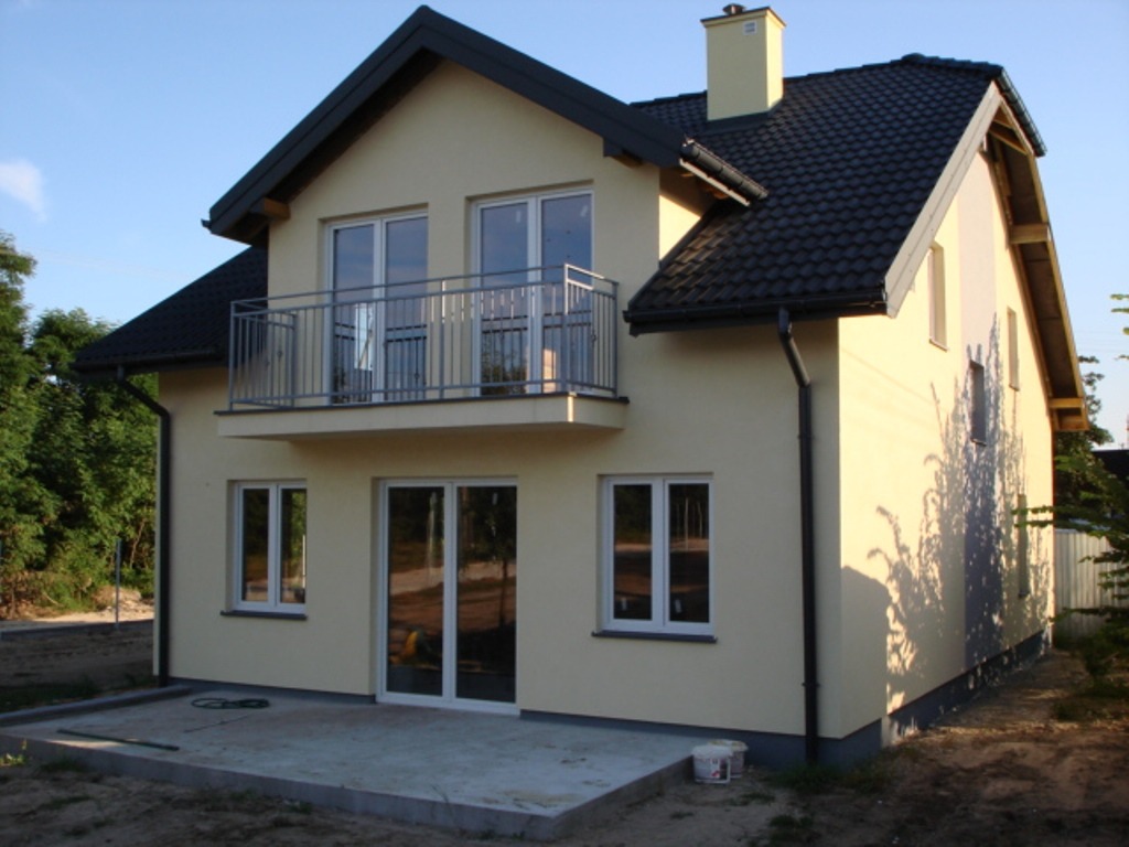 Dwukondygnacyjny dom z balkonem i żółtą elewacją w słoneczny dzień, widoczne świeże fundamenty tarasu.