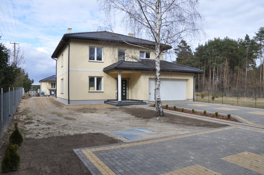 Dwupiętrowy dom z beżową elewacją i ciemnym dachem, z podjazdem wyłożonym kostką brukową i młodymi drzewkami wzdłuż ścieżki, widoczny drugi dom w tle.