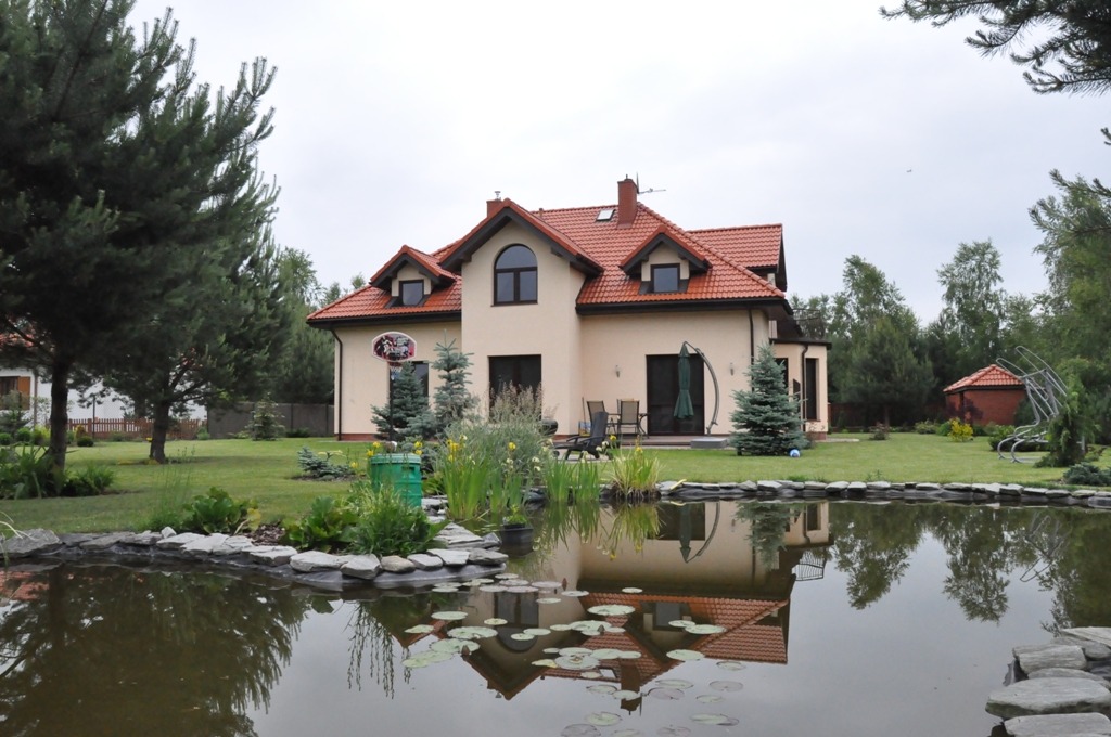 Dwukondygnacyjny dom z czerwonym dachem, odbijający się w stawie z liliami wodnymi; widoczny zadbany ogród z iglakami i elementami małej architektury.
