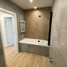 Ivan Kozar - Nowoczesna łazienka po remoncie z płytkami imitującymi drewno na ścianie za wanną i podłodze, szklany parawan nawannowy z czarnymi elementami, grzejnik drabinkowy i szare płytki na pozostałych...